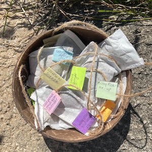 May include: A brown wooden bowl filled with small white tea bags tied with twine and labeled with handwritten notes. The notes include "Rose petals lemon balm lavender", "Sniffle N Sneeze", "Minty Pick-me-up", "Cornice Ginger", "Dandelion", "Chamomile", and "Almond Balm Lavender".