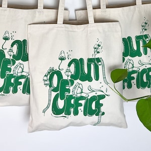 May include: Three white canvas tote bags with green text and illustrations. The bags are hanging on a copper wire rack. The text on the bags reads "Out of Office".