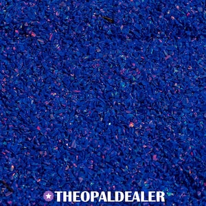 May include: Close-up of a pile of crushed, deep blue opal. The opal pieces have iridescent flashes of pink, orange, and green. The text "THEOPALDEALER" is at the bottom of the image.