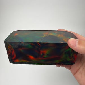 May include: A rectangular, iridescent gemstone with a polished surface. The stone displays a play of colors, including red, green, and orange, against a dark background. The gemstone is held in a hand.