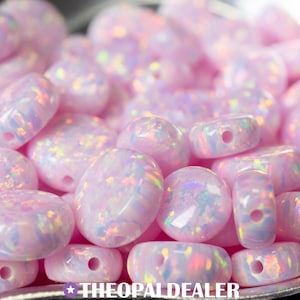 May include: Close-up of a collection of pink opal beads with iridescent flashes. The beads are in various shapes, including round, oval, and barrel, each with a drilled hole. The text "THEOPALDEALER" is at the bottom.