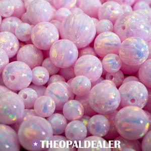 May include: A pile of pink opal beads in various sizes. The beads have a pearlescent sheen with iridescent flashes of color. The text "THEOPALDEALER" is at the bottom of the image.