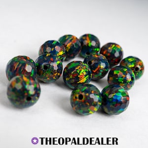 May include: A collection of faceted, dark blue opal beads with vibrant flashes of green, yellow, and orange. Each bead has a drilled hole and a unique, colorful pattern. The Opal Dealer logo is at the bottom.
