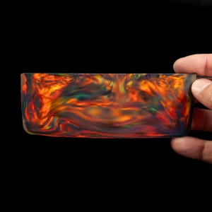 May include: A rectangular block of polished resin featuring a swirling, marbled pattern of vibrant orange, deep red, and dark black tones with iridescent flashes of green and yellow throughout the smooth, semi-translucent material surface.