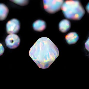 May include: A collection of iridescent, faceted opal gemstones against a black background. The gemstones have a diamond-like shape and reflect a spectrum of colors, including blue, pink, and green. Some are round, others are more angular.