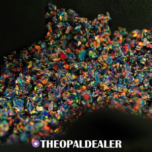 Puede incluir: Una colección de pequeños fragmentos de ópalo triturados e irregulares que muestran un vibrante juego de colores con destellos iridiscentes de rojo, naranja, amarillo, verde y azul sobre una base oscura. El texto THEOPALDEALER aparece abajo.