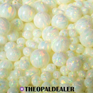 May include: A collection of iridescent white opal beads in various sizes. The beads have a pearly, opalescent sheen with hints of rainbow colors. The beads are scattered, creating a textured, eye-catching display. The text "THEOPALDEALER" is at the bottom.