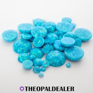 May include: A collection of blue opal cabochons in various sizes, with a mix of round and oval shapes. The opals have a vibrant, swirling pattern of blue and green hues. The text "THEOPALDEALER" is at the bottom.