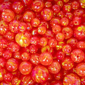 May include: A close-up of numerous red and orange beads with a glossy, iridescent finish. The beads, of varying sizes, display hints of yellow and gold. Each bead is pierced, indicating their use in jewellery making or other craft projects.