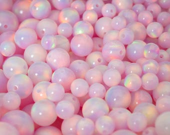 May include: A collection of pink and iridescent opal beads of varying sizes. The beads are round with a glossy finish and have a subtle play of color. The text "THEOPALDEALER" is at the bottom.