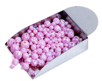May include: A collection of pink opal beads of varying sizes spills from a silver metal container. The beads have a pearlescent, iridescent quality, with a mix of solid and patterned surfaces. The container has a curved shape and a hinged lid.