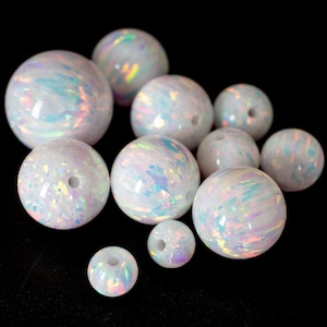 May include: A collection of white opal beads with iridescent swirls and a glossy finish. The beads vary in size and have a hole drilled through the center, suggesting they are for jewelry making or crafting. The background is black.
