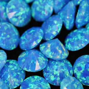 May include: A selection of oval-cut blue opal gemstones. The opals display a shimmering, iridescent effect with hints of green and yellow. The gemstones are arranged against a dark backdrop, highlighting their colour and sparkle.