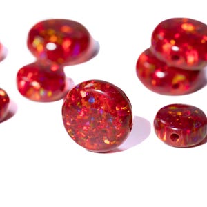 Könnte beinhalten: Nahaufnahme von mehreren roten Opalperlen mit glänzendem Finish. Die Perlen haben ein gesprenkeltes Aussehen mit Farbtupfern, darunter Orange, Gelb und Blau. Eine Perle hat ein Loch. Die Perlen sind auf weißem Hintergrund.