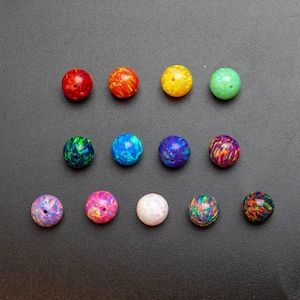 May include: A collection of thirteen spherical beads arranged in three rows. Each bead features a distinct iridescent finish with vibrant color patterns including red, orange, yellow, green, blue, purple, pink, white, and black with multi-colored flecks.