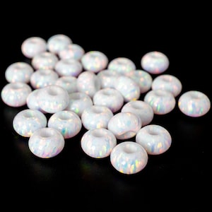 May include: A collection of white, donut-shaped beads with iridescent, opal-like surfaces. The beads are scattered against a black background, showcasing their unique texture and color variations. These beads are ideal for jewelry making.
