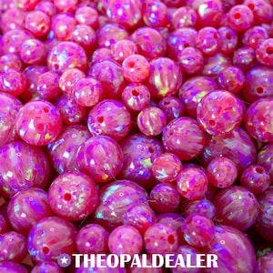 Puede incluir: Un conjunto de cuentas de ópalo rosas y moradas de varios tamaños. Las cuentas tienen un acabado brillante e iridiscente, con patrones internos visibles. El texto "THEOPALDEALER" está en la parte inferior de la imagen.