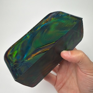 May include: A rectangular, iridescent object with a dark, textured exterior and a swirling, multi-colored interior. The colors include green, blue, and orange, creating a vibrant, oil-slick effect. The object is held in a hand.