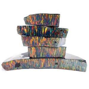 May include: A stack of six rough-cut opal stones. The opals display a vibrant play-of-color, with streaks of yellow, orange, green, and blue against a dark background. The stones are irregularly shaped, showcasing their natural, raw texture.