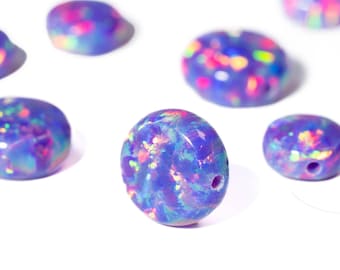 May include: A collection of blue opal beads with iridescent flecks of pink, yellow, and green. The beads are oval and round, with a polished surface and a small hole for stringing. The beads are scattered on a white background.