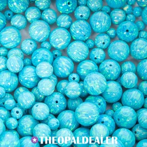 May include: A collection of round, blue opal beads in various sizes. The beads have a swirling, iridescent pattern and are drilled for stringing. The text "THEOPALDEALER" is at the bottom of the image.