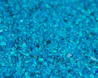 May include: A collection of small, crushed bright blue opal fragments with a reflective, iridescent finish. The texture is granular and uneven, featuring varying shades of cyan and azure. The text THEOPALDEALER is printed in white at the bottom.
