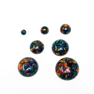 May include: Seven polished opal gemstones in a variety of sizes. The opals are round and display a mix of colours, including blue, green, orange, and red, against a dark base. The largest opals are at the bottom.