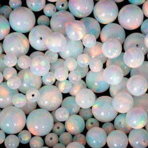 May include: A collection of iridescent, white opal beads in various sizes. The beads have a pearlescent sheen with hints of blue, green, and pink. Some beads have holes for stringing. The background is black.