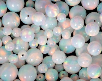 May include: A collection of iridescent, white opal beads in various sizes. The beads have a pearlescent sheen with hints of blue, green, and pink. Some beads have holes for stringing. The background is black.
