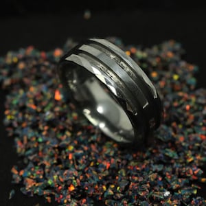 May include: A silver ring with a black inlay and a faceted design, set against a backdrop of colourful, iridescent fragments. The ring's surface reflects light, highlighting its intricate details. The ring is a piece of jewellery.