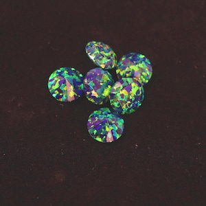 Op de afbeelding: Een verzameling van zes ronde, gefacetteerde edelstenen. De edelstenen tonen een levendige mix van kleuren, waaronder blauw, groen en paars. De edelstenen liggen op een donkerbruine achtergrond.