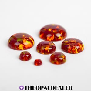 May include: A collection of cabochon-cut opals in various sizes. The opals are a deep red color with flashes of yellow and green. The opals are displayed on a white surface. The text "THEOPALDEALER" is at the bottom.