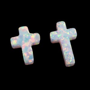May include: Two white opal crosses with iridescent flecks. The crosses are set against a black background. The opal material has a pearly, shimmering quality, reflecting various colors.