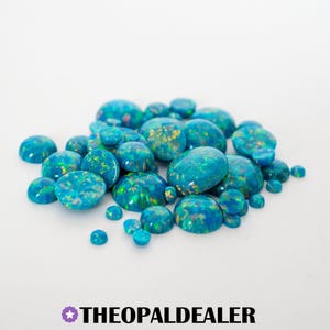 May include: A collection of polished opal gemstones in various sizes and shapes. The opals display a vibrant mix of blue, green, and gold colors, with a glossy, domed surface. The text "THEOPALDEALER" is at the bottom.