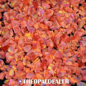 May include: Close-up of a pile of crushed, iridescent opal gemstones. The opals are primarily shades of orange and pink, with flashes of yellow and purple. The text "THEOPALDEALER" is at the bottom.