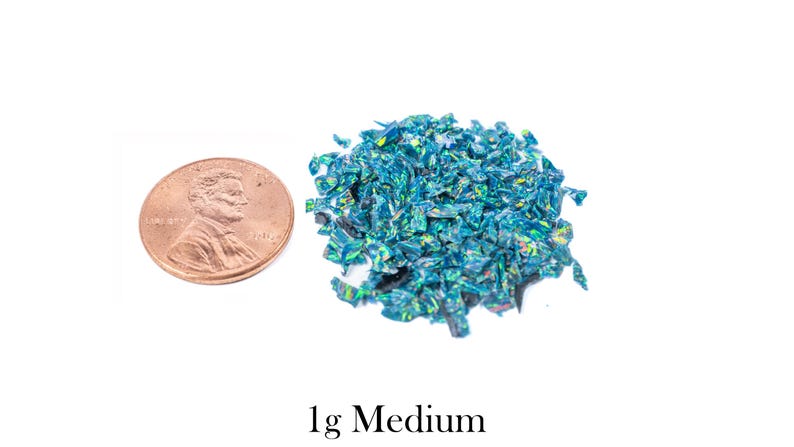 Puede incluir: Un mont&oacute;n de fragmentos de vidrio triturado azul-verdoso iridiscente junto a una moneda. El texto "1g Medium" est&aacute; debajo. Los fragmentos tienen un aspecto reflectante y brillante.