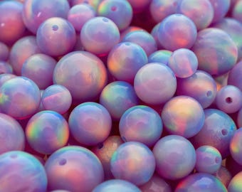 May include: A close-up of a collection of iridescent, round beads in shades of purple, pink, and blue. The beads vary in size and have a glossy finish. The text "THEOPALDEALER" is at the bottom of the image.