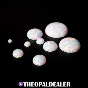 May include: A collection of white opal cabochons with iridescent flashes of color. The gemstones are in various sizes, with a smooth, rounded shape. The opals are displayed against a black background. The text "THEOPALDEALER" is at the bottom.
