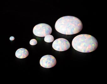 May include: A collection of white opal cabochons with iridescent flashes of color. The gemstones are in various sizes, with a smooth, rounded shape. The opals are displayed against a black background. The text "THEOPALDEALER" is at the bottom.