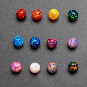 May include: A collection of twelve spherical beads arranged in three rows of four. Each bead features a distinct color pattern, including red, orange, yellow, green, blue, purple, pink, and white, with iridescent flecks throughout the surfaces.