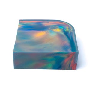 May include: A rectangular soap bar with a rounded corner. The soap has a swirling, iridescent pattern of blue, green, yellow, and red colors. The bar is opaque and appears to be made of a solid material.
