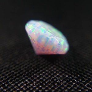 May include: A faceted, oval-shaped opal gemstone. The opal displays a play of color with flashes of green, blue, and pink hues. The gemstone is set against a dark background, highlighting its iridescent qualities.