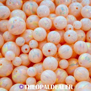 May include: A close-up of a collection of round, opalescent beads in varying sizes. The beads are mainly peach and white, with iridescent flashes of colour. The beads have holes for stringing and are closely packed together.