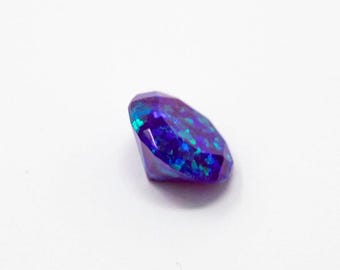 May include: A faceted, oval gemstone with a deep blue base and iridescent teal and purple flashes. The gem is set against a plain white background, highlighting its brilliant cut and colour variations.