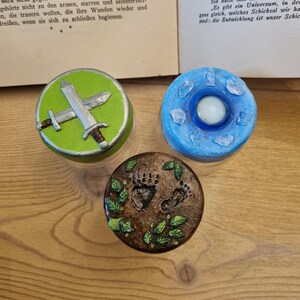 May include: Three small glass jars with decorative lids. The lids are painted with different designs: a green lid with a silver sword, a blue lid with a white circle and a brown lid with two footprints and green leaves.
