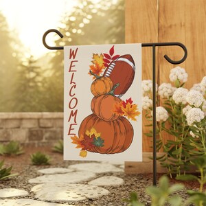 May include: A welcome garden flag with a stack of pumpkins and a football. The pumpkins are orange and brown with fall leaves. The football is brown and white. The flag is white with the word "WELCOME" in red.