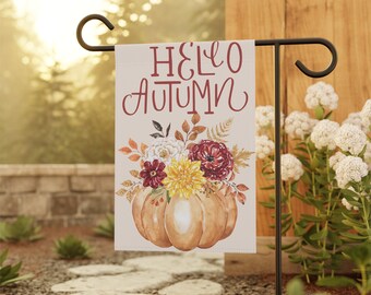 May include: A garden flag with a white background and the text "Hello Autumn" in red. The flag features a watercolor illustration of a pumpkin with fall flowers.
