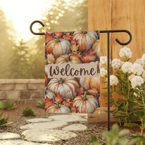 May include: A garden flag featuring a watercolor design of pumpkins and fall leaves. The flag has the word "Welcome" printed on it. The flag is displayed on a black metal stand in a garden setting.