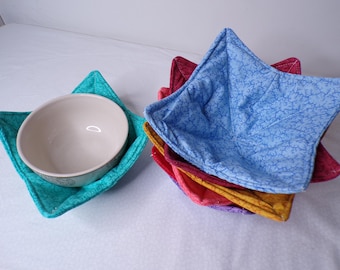 May include: A set of quilted fabric bowl cosies in various colours including teal, light blue, yellow, pink, and purple. One teal bowl cosy holds a plain beige ceramic bowl, while the others are stacked together in a nested arrangement.