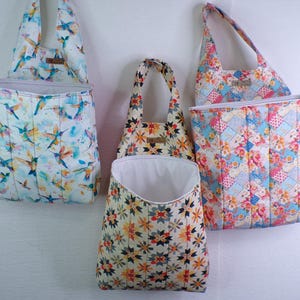 May include: Three hanging fabric storage organizers with quilted vertical stitching. The left bag features a hummingbird print, the center bag has a geometric star pattern, and the right bag displays a floral patchwork design with white interiors.